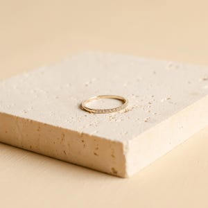 May include: A delicate gold ring with small, clear gemstones set along the band. The ring is displayed on a textured, off-white surface against a soft, neutral background. The ring's design is simple and elegant.
