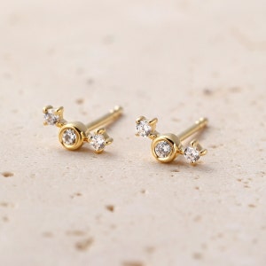 May include: A pair of gold stud earrings. Each earring has a central, round, clear gemstone in a gold bezel, with two smaller clear gemstones extending outwards. The earrings are presented on a light-coloured surface.