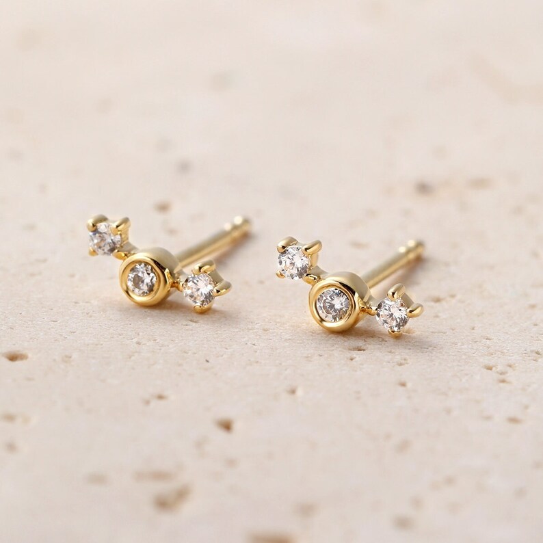 May include: A pair of gold stud earrings. Each earring has a central, round, clear gemstone in a gold bezel, with two smaller clear gemstones extending outwards. The earrings are presented on a light-coloured surface.