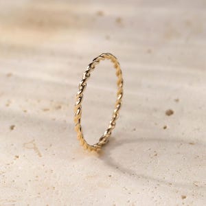 May include: A gold-colored ring with a twisted rope design. The ring is a thin band, likely made of metal, and sits on a neutral-toned surface. The ring's texture is visible, with the twisted pattern creating shadows and highlights.