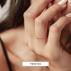 K&ouml;nnte beinhalten: Ein zarter Goldring mit einem gedrehten Band und einem kleinen Diamanten als Akzent. Der Ring wird an einem Finger getragen.