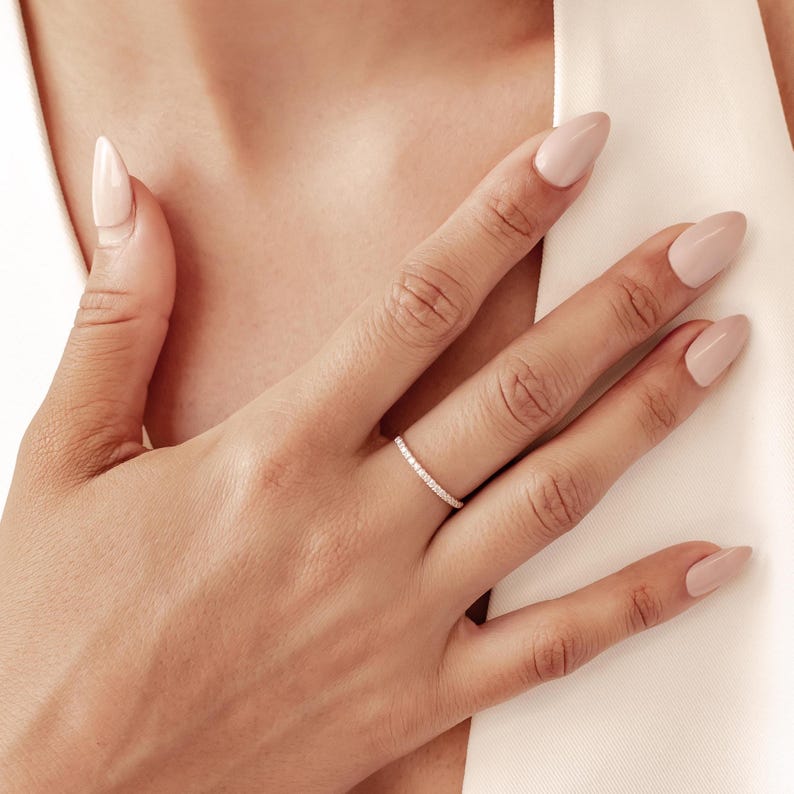 May include: A delicate, thin ring with a band of small, sparkling stones. The ring is set on a hand with manicured nails, suggesting a focus on jewelry and accessories. The ring's design is simple and elegant, suitable for everyday wear.