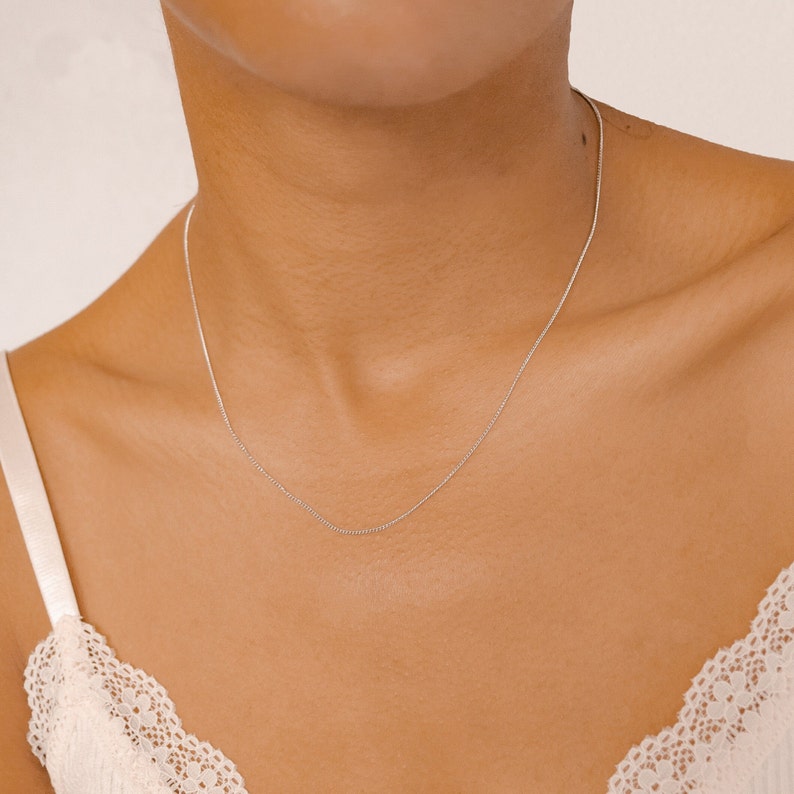 May include: A simple silver chain necklace worn on a person's neck. The chain is thin and delicate, making it a popular choice for everyday wear.