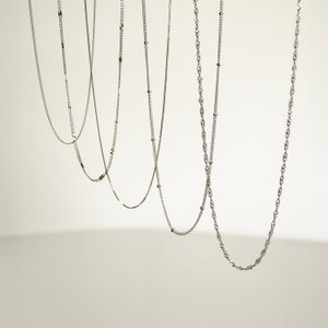 May include: Five silver chains of different styles and lengths hanging against a white background.