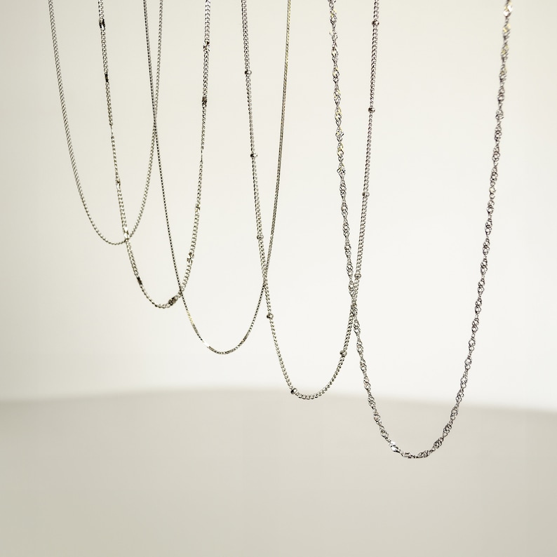 May include: Five silver chains of different styles and lengths hanging against a white background.