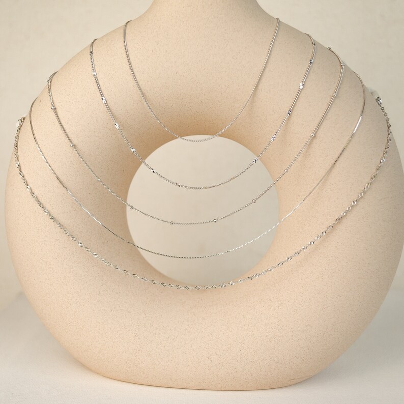 May include: A set of seven silver chains of varying lengths and styles displayed on a beige ceramic stand.