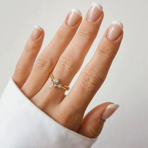 May include: A gold-colored ring with a V-shaped design, adorned with small, clear gemstones. The ring is on a person's finger, with a white shirt sleeve in the background. The nails are painted with a French manicure.