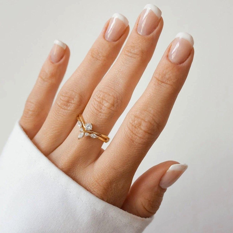 May include: A gold-colored ring with a V-shaped design, adorned with small, clear gemstones. The ring is on a person's finger, with a white shirt sleeve in the background. The nails are painted with a French manicure.
