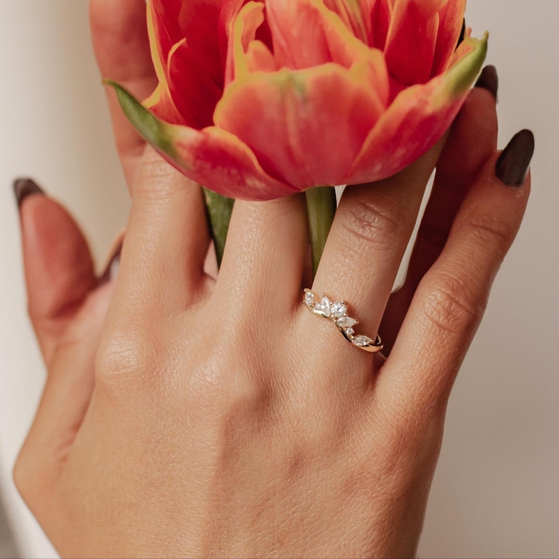 May include: A gold ring with a floral design featuring multiple small, clear gemstones. The ring is worn on a finger, and a red and yellow flower is held in the same hand. The ring's design is delicate and elegant.