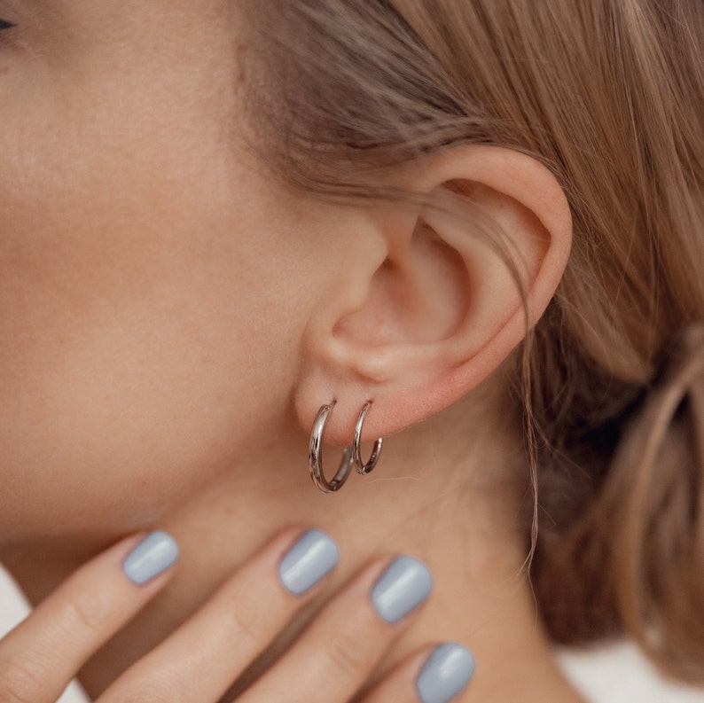 May include: A pair of silver hoop earrings worn in the left ear. The earrings are simple and understated, with a smooth, polished finish.