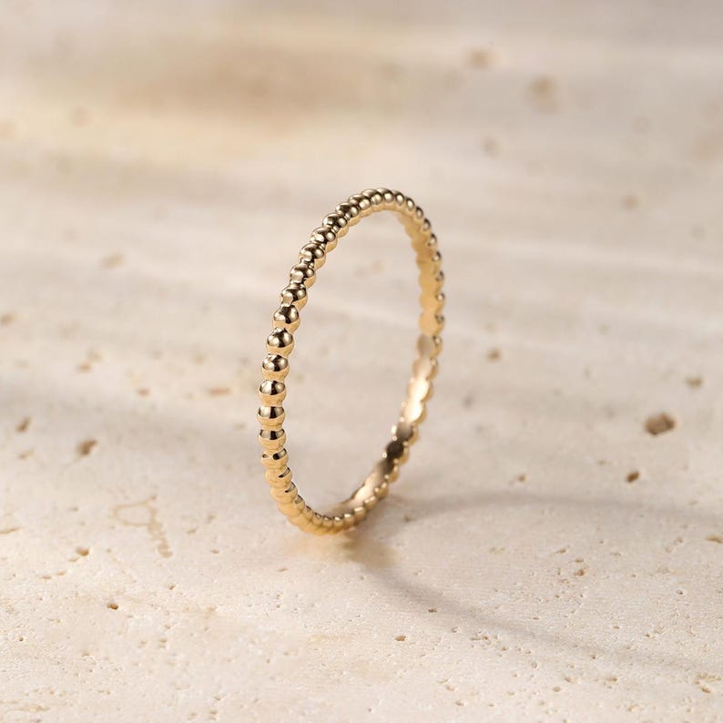 May include: A delicate gold ring with a beaded design. The ring features a series of small, evenly spaced beads around the band, creating a textured and elegant appearance. The ring is set against a neutral background.