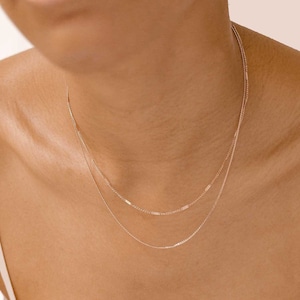 Könnte beinhalten: Zwei zarte goldene Ketten, die auf dem Hals einer Person übereinander liegen. Die Ketten sind dünn und haben einen dezenten Glanz.
