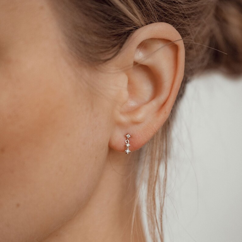 Puede incluir: Primer plano de una oreja con un peque&ntilde;o y delicado pendiente de plata con una cadena y tres peque&ntilde;os diamantes.