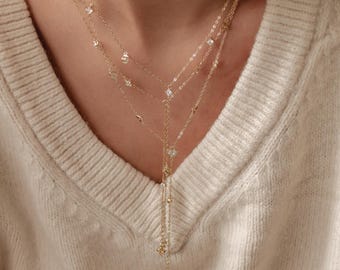 Könnte beinhalten: Eine mehrschichtige Goldkette mit filigranen Ketten und funkelnden Akzenten. Die Halskette hat mehrere Stränge unterschiedlicher Längen, die mit kleinen, klaren Edelsteinen und geometrischen Formen verziert sind. Das Design beinhaltet einen Y-förmigen Tropfen mit einem hängenden Anhänger.