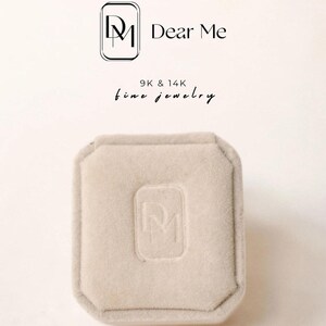 May include: A square, beige jewelry box with a soft, velvety texture. The box has a recessed logo on the top and is set against a light background. The text "Dear Me" and "9K & 14K fine jewelry" are displayed above the box.