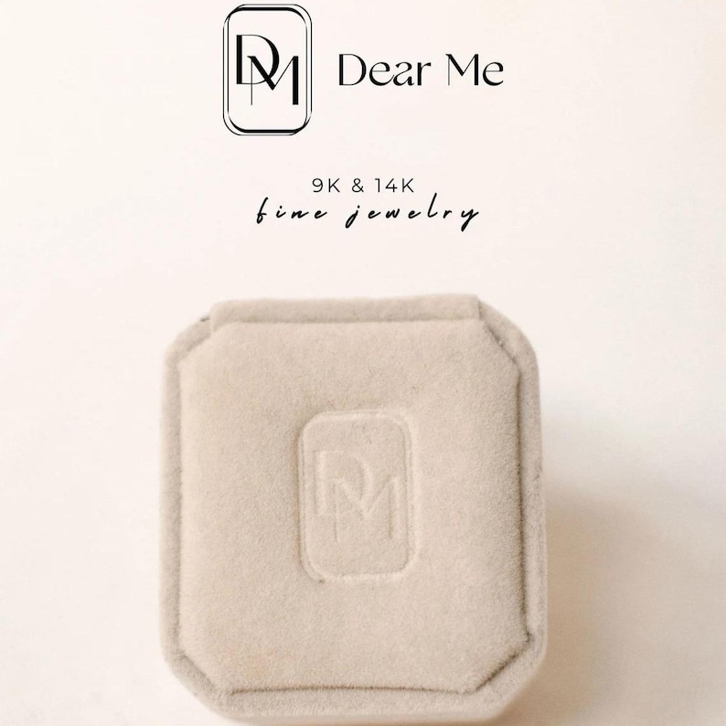 May include: A square, beige jewelry box with a soft, velvety texture. The box has a recessed logo on the top and is set against a light background. The text "Dear Me" and "9K & 14K fine jewelry" are displayed above the box.