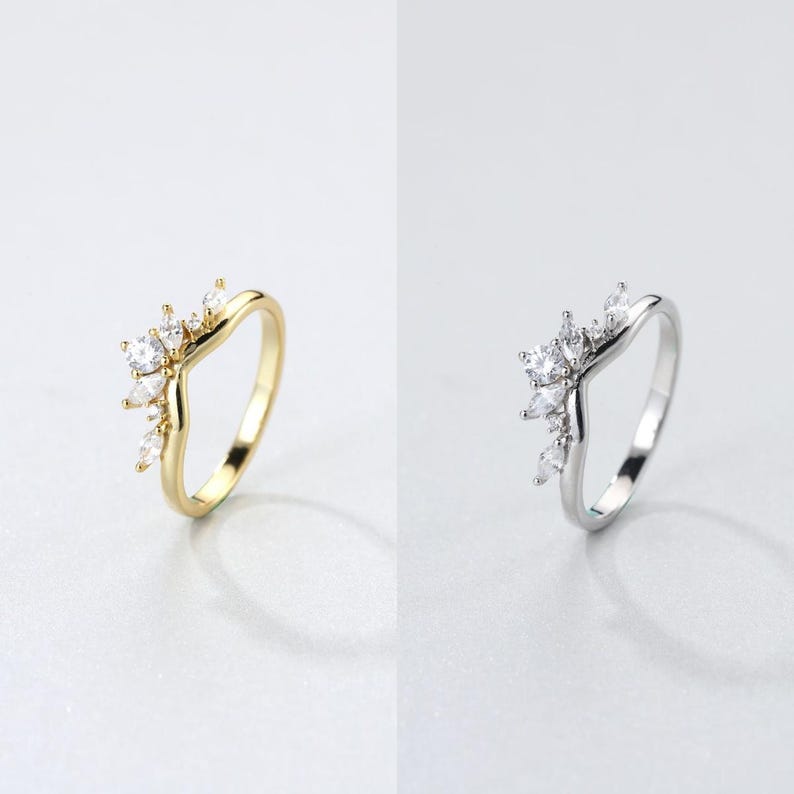 May include: Two rings, one gold and one silver, each with a V-shaped band. The gold ring features a cluster of clear, sparkling stones at the top. The silver ring has a similar design with clear stones.