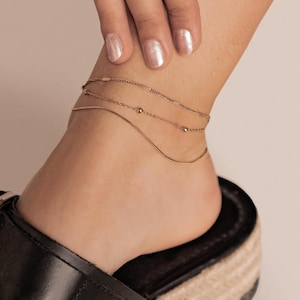 May include: A close-up of three delicate gold anklets. The top anklet features small rectangular bars, the middle one has tiny beads, and the bottom one is a simple chain. The anklets are worn on an ankle with a black shoe in the foreground.