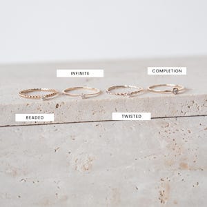 K&ouml;nnte beinhalten: F&uuml;nf goldene Ringe mit kleinen Diamanten, die in die Band eingefasst sind. Die Ringe sind in einer Reihe auf einer hellbeigen, strukturierten Oberfl&auml;che angeordnet. Die Ringe sind mit "Beaded", "Infinite", "Twisted", "Completion" beschriftet.