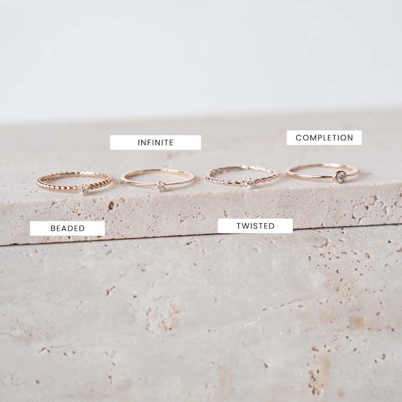 K&ouml;nnte beinhalten: F&uuml;nf goldene Ringe mit kleinen Diamanten, die in die Band eingefasst sind. Die Ringe sind in einer Reihe auf einer hellbeigen, strukturierten Oberfl&auml;che angeordnet. Die Ringe sind mit "Beaded", "Infinite", "Twisted", "Completion" beschriftet.