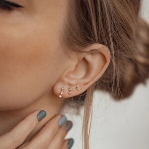 Puede incluir: Primer plano de la oreja de una persona con tres peque&ntilde;os pendientes de oro con peque&ntilde;os diamantes. Los pendientes est&aacute;n en los piercings del l&oacute;bulo, el h&eacute;lix y el l&oacute;bulo superior.
