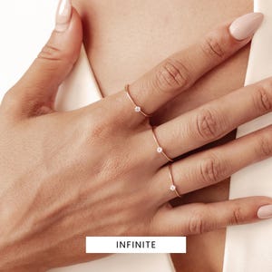 K&ouml;nnte beinhalten: Drei zarte goldene Ringe, jeder mit einem kleinen Diamanten besetzt. Die Ringe werden an den Fingern einer Hand getragen. Das Wort "INFINITE" ist unter der Hand gedruckt.