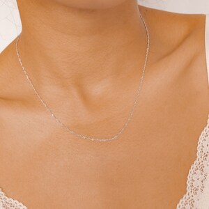 May include: A delicate silver chain necklace worn on a person's neck. The chain is thin and has a subtle shimmer.