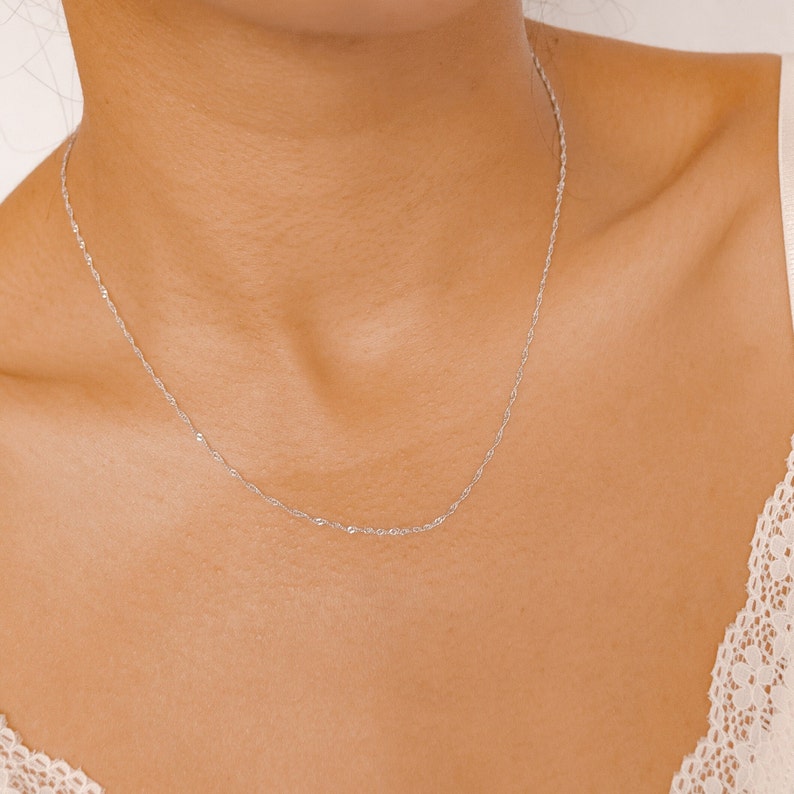 May include: A delicate silver chain necklace worn on a person's neck. The chain is thin and has a subtle shimmer.