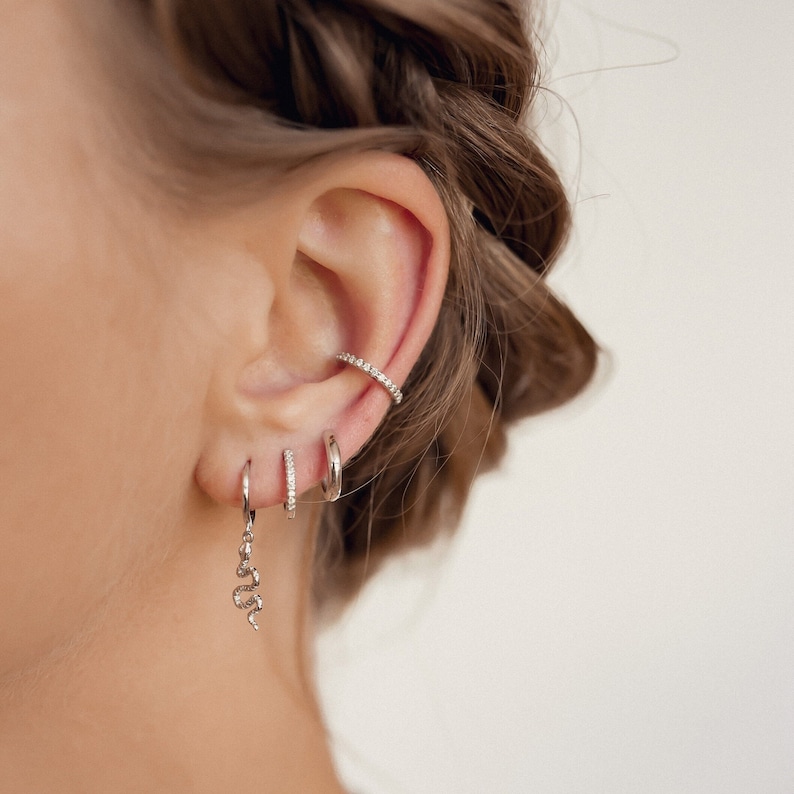 Puede incluir: Primer plano de la oreja de una persona con varios pendientes de plata. Los pendientes incluyen un aro peque&ntilde;o con un dije de serpiente, un aro m&aacute;s peque&ntilde;o con diamantes pav&eacute; y un aro m&aacute;s grande con diamantes pav&eacute;.