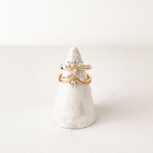 May include: Two gold-colored rings with clear gemstones are displayed on a white, cone-shaped stand. The rings have a delicate, crown-like design, with the gemstones arranged in a symmetrical pattern. The stand is textured and has a matte finish.