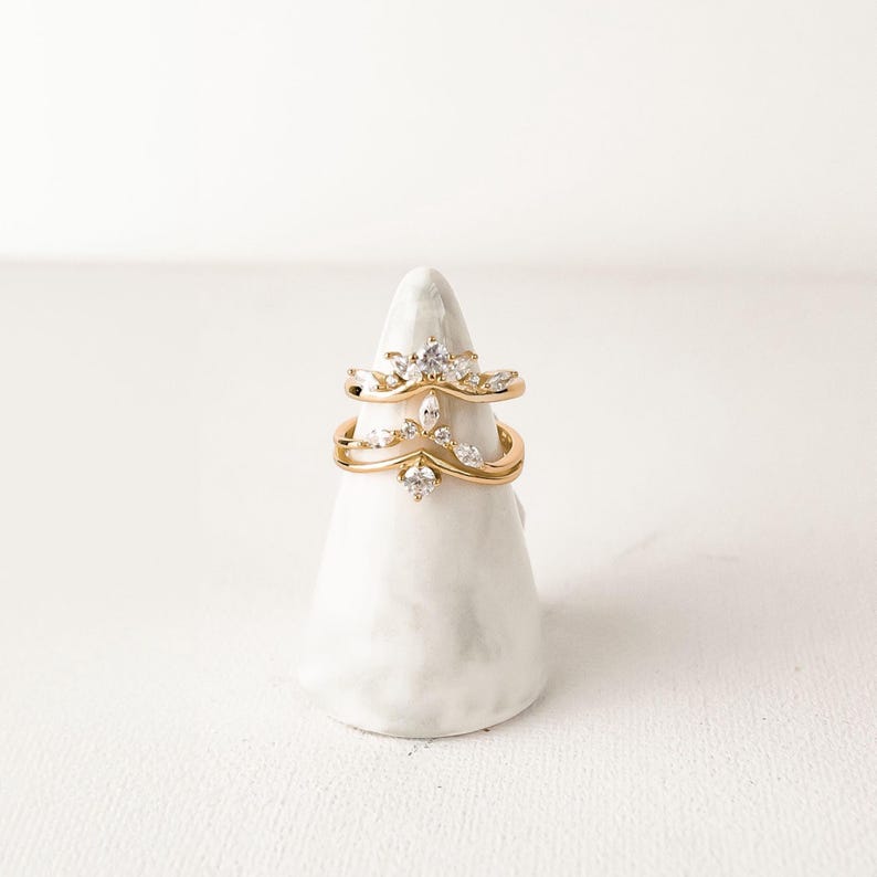 May include: Two gold-colored rings with clear gemstones are displayed on a white, cone-shaped stand. The rings have a delicate, crown-like design, with the gemstones arranged in a symmetrical pattern. The stand is textured and has a matte finish.