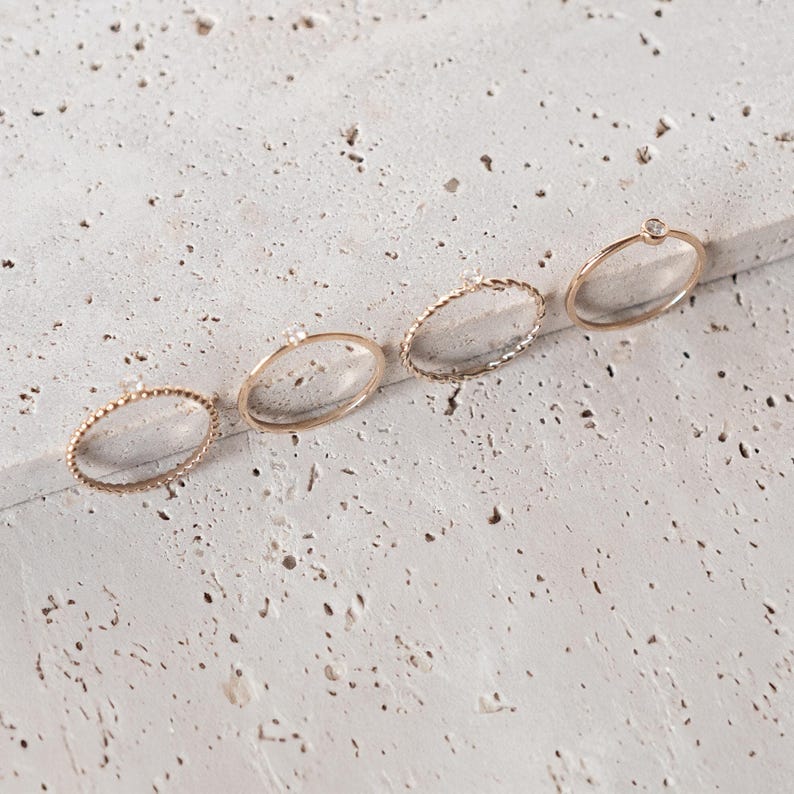K&ouml;nnte beinhalten: F&uuml;nf goldene Ringe mit kleinen Diamanten auf einer wei&szlig;en, strukturierten Oberfl&auml;che. Die Ringe sind in einer Reihe von links nach rechts angeordnet, wobei der Ring mit dem gr&ouml;&szlig;ten Diamanten rechts liegt.