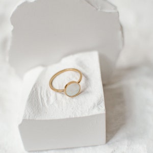 K&ouml;nnte beinhalten: Ein goldener Ring mit einem wei&szlig;en runden Stein in der Mitte. Der Ring liegt auf einer wei&szlig;en Oberfl&auml;che.