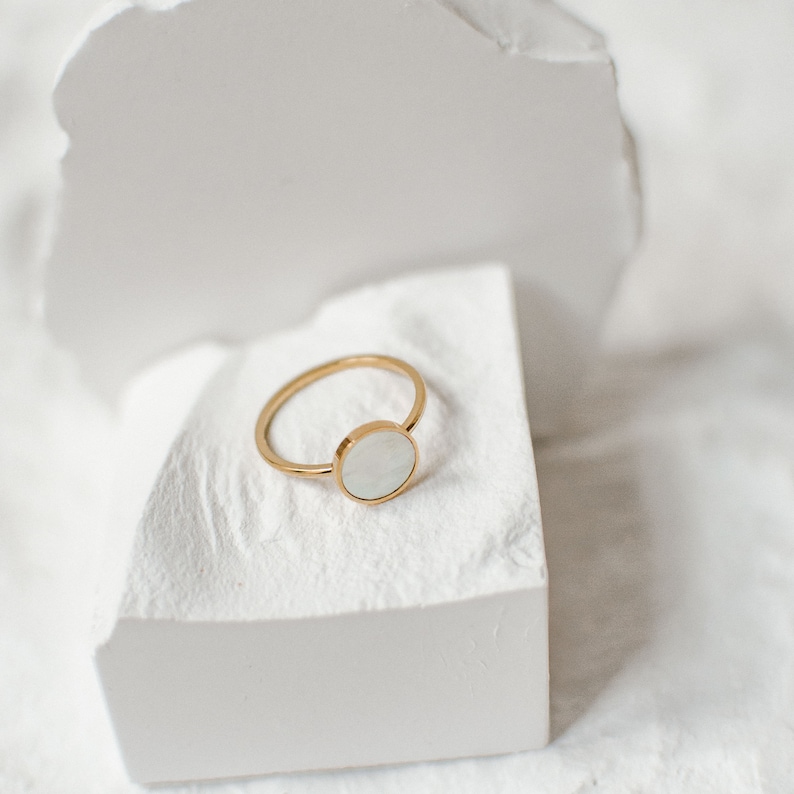 K&ouml;nnte beinhalten: Ein goldener Ring mit einem wei&szlig;en runden Stein in der Mitte. Der Ring liegt auf einer wei&szlig;en Oberfl&auml;che.