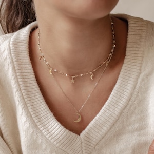 Può includere: Una collana dorata a due strati con ciondoli celesti. Lo strato superiore ha piccole mezzelune e una stella. Lo strato inferiore ha un ciondolo a mezzaluna più grande. La collana è indossata sopra un maglione color crema.