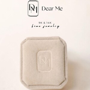 May include: A light beige jewelry box with a soft, velvety texture. The box has a rectangular shape with a slightly octagonal lid. The embossed logo of "Dear Me" is visible on the lid. The text "9K & 14K fine jewelry" is also visible.