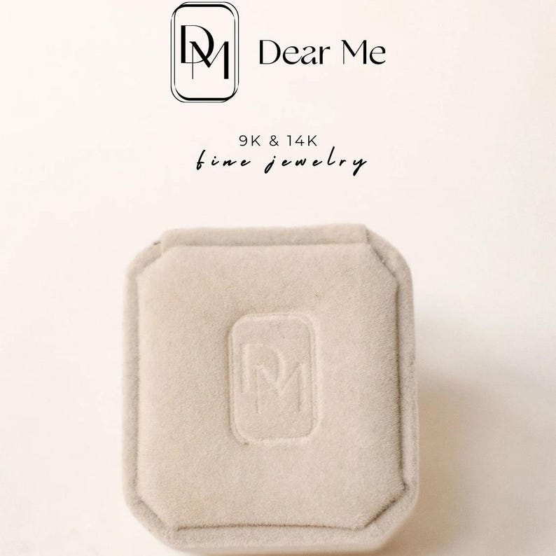May include: A light beige jewelry box with a soft, velvety texture. The box has a rectangular shape with a slightly octagonal lid. The embossed logo of "Dear Me" is visible on the lid. The text "9K & 14K fine jewelry" is also visible.