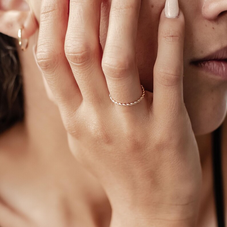 May include: A delicate, thin gold ring with a twisted rope design. The ring is worn on a finger, and the image is a close-up, highlighting the ring's texture and shine. The ring is a simple yet elegant piece of jewelry.