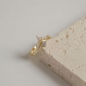 May include: A gold-colored ring with a unique design. The ring features a series of small, clear gemstones and a larger, central stone. The band has a double-layered design. The ring is displayed on a textured, light-colored surface.
