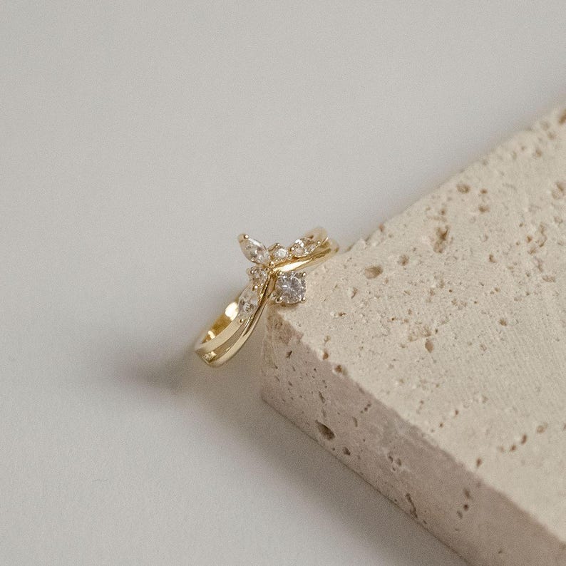 May include: A gold-colored ring with a unique design. The ring features a series of small, clear gemstones and a larger, central stone. The band has a double-layered design. The ring is displayed on a textured, light-colored surface.