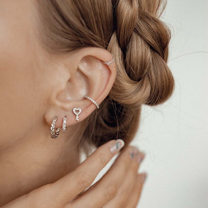 May include: A close-up of a person's ear with multiple silver earrings. The earrings include a small key with a heart-shaped hole, a small hoop, and a larger hoop with a braided design.