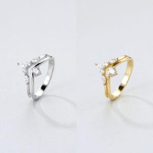 May include: Two rings, one silver and one gold, are displayed. Each ring features a V-shaped design with a cluster of small, clear gemstones. The silver ring has a double band, while the gold ring has a single band.