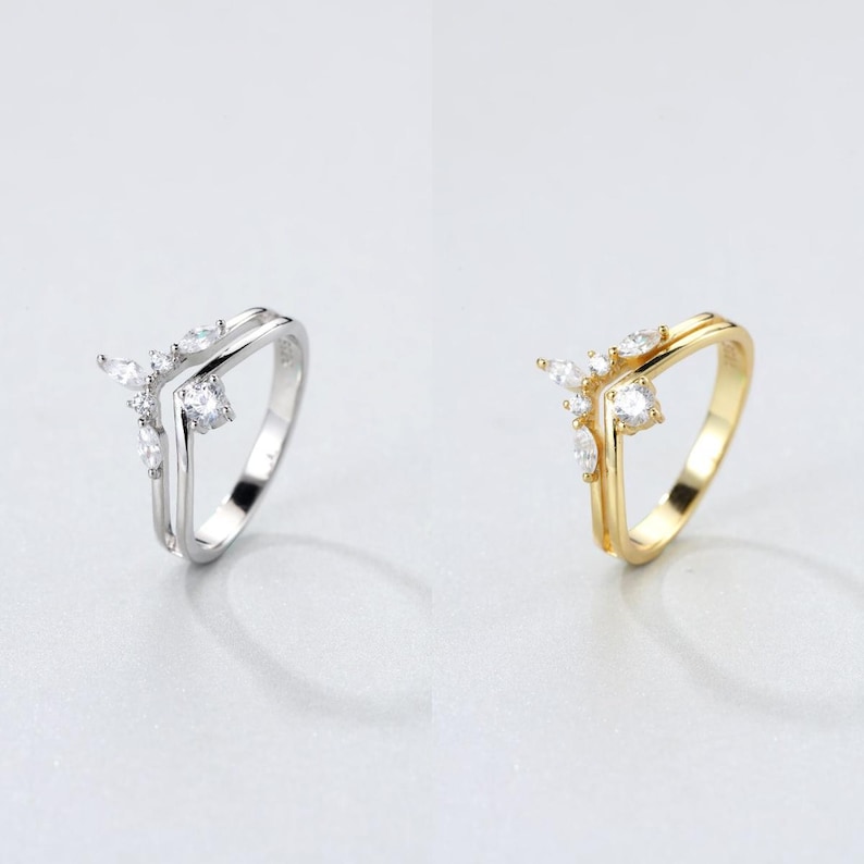 May include: Two rings, one silver and one gold, are displayed. Each ring features a V-shaped design with a cluster of small, clear gemstones. The silver ring has a double band, while the gold ring has a single band.
