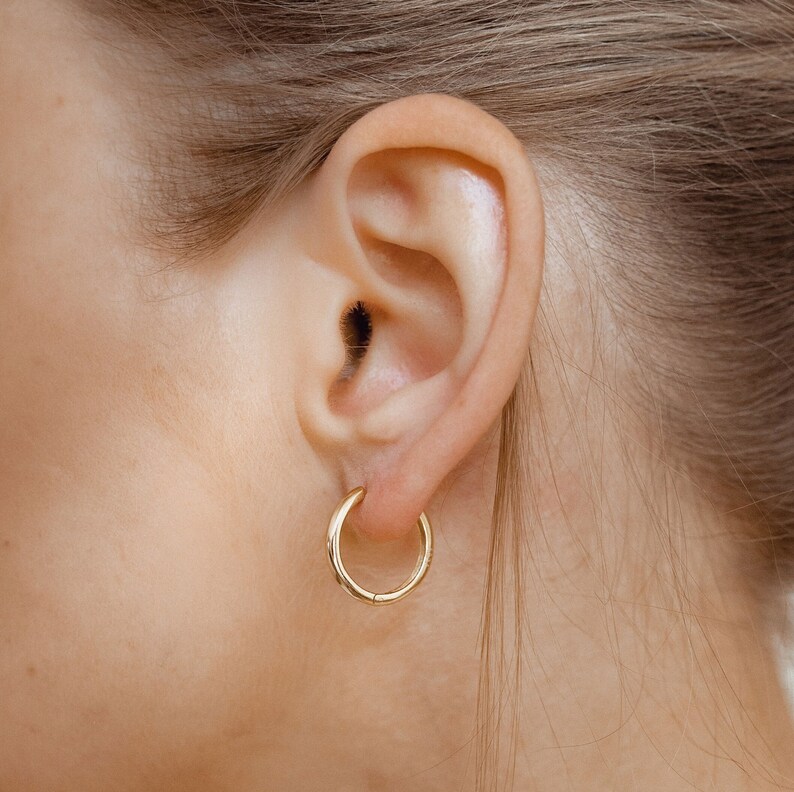 May include: A close-up of a person's ear with a gold hoop earring. The earring is simple and elegant, with a smooth, polished finish.