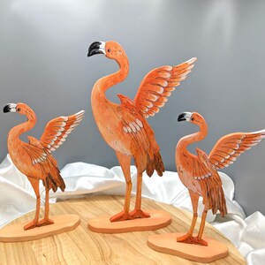 Può includere: Tre sculture di fenicotteri arancioni con le ali spiegate. Gli uccelli hanno becchi neri e sono montati su basi di legno. Le sculture sono dipinte a mano con dettagliati motivi di piume, creando un tema tropicale decorativo.