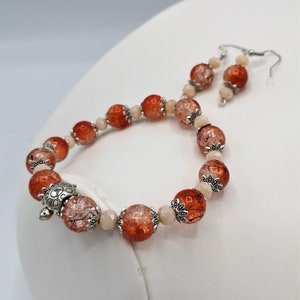 Op de afbeelding: Een sieradenset bestaande uit een armband en bijpassende oorbellen. De armband heeft grote, oranje-rode craquelé kralen, kleinere lichtroze kralen en zilveren accenten. De oorbellen weerspiegelen het ontwerp met vergelijkbare kralen en zilveren haken. Een zilveren schildpadbedel siert de armband.