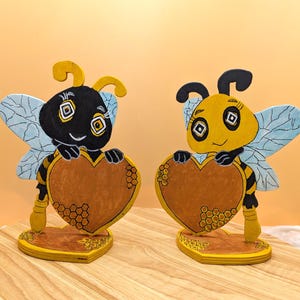 Könnte beinhalten: Zwei Holz-Bienenfiguren, eine schwarze und eine gelbe, die jeweils ein braunes Herz halten. Die Bienen haben blaue Flügel und stehen auf einem gelben Sockel. Der Hintergrund ist hellorange.