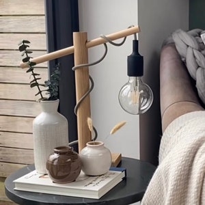 Op de afbeelding: Een houten lamp met een grijs snoer en een zwarte fitting. De lamp staat op een bijzettafel met een boek en drie vazen, twee witte en een bruine.