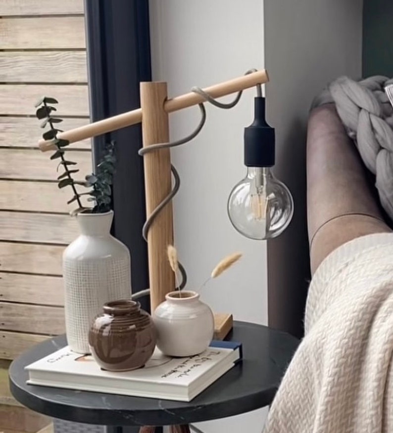 Op de afbeelding: Een houten lamp met een grijs snoer en een zwarte fitting. De lamp staat op een bijzettafel met een boek en drie vazen, twee witte en een bruine.
