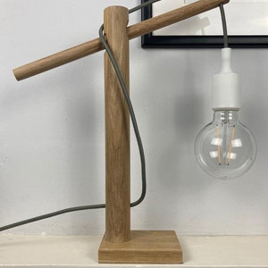 Op de afbeelding: Een houten tafellamp met een grijs snoer en een witte lamp. De lamp heeft een lange, horizontale arm die zich uitstrekt vanaf de basis.